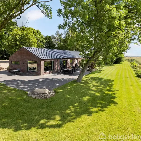 En moderne enfamilieshus i røde mursten står omgivet af grønne plæner og høje træer på grunden. Dagslys reflekteres i store vinduer, og området er landligt og frodigt.