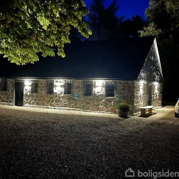 En lang, lav stensbygning oplyst af udendørs lamper, der skaber en hyggelig atmosfære. Bygningen står i en gruset gårdsplads med en hvid bil parkeret til højre.