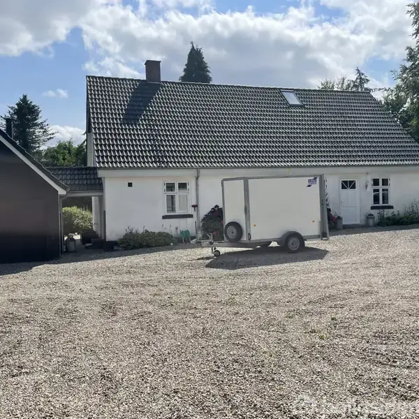 Hvidt hus med gråt tag står stille i en gårdsplads med grus. En trailer er parkeret foran, og der er en garage til venstre omkranset af træer og buske.