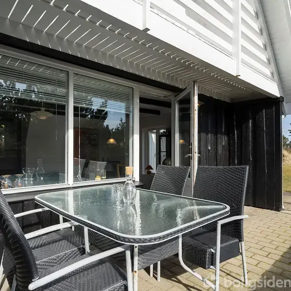 Et glasbord med sorte flettestole står på en terrasse af fliser. I baggrunden ses et sort hus med store vinduer og natur omgivet af træer.