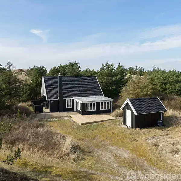 Et sort træhus står stille på en solbelyst grund omgivet af buske og træer. Ved siden af ligger et mindre skur. Alt er placeret i et naturskønt område.