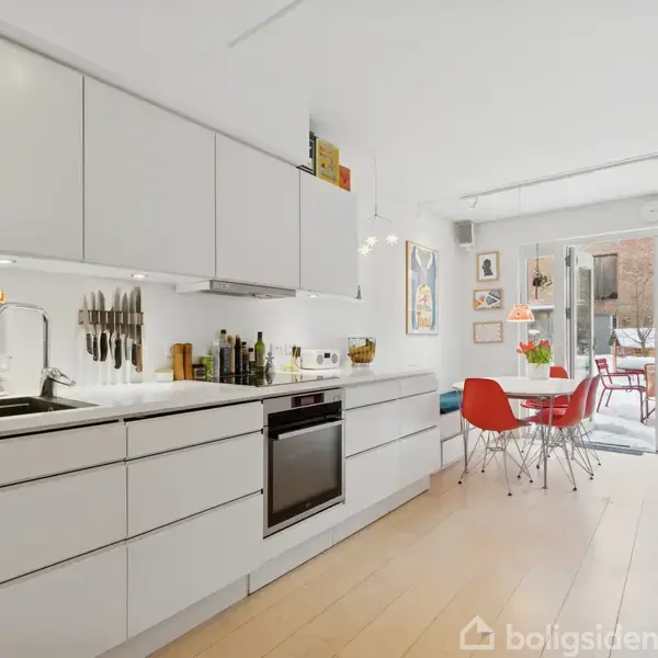 Et moderne køkken med hvide skabe inklusiv en ovn, placeret i et lyst rum. Til højre ses et spisebord med røde stole, der fører ud til en terrasse.