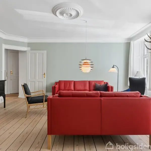 En rød sofa står centralt i en stue med trægulv og grønne vægge. En moderne lampe hænger over sofaen. En sort skænk, en stol og et hjortegevir pryder rummet.