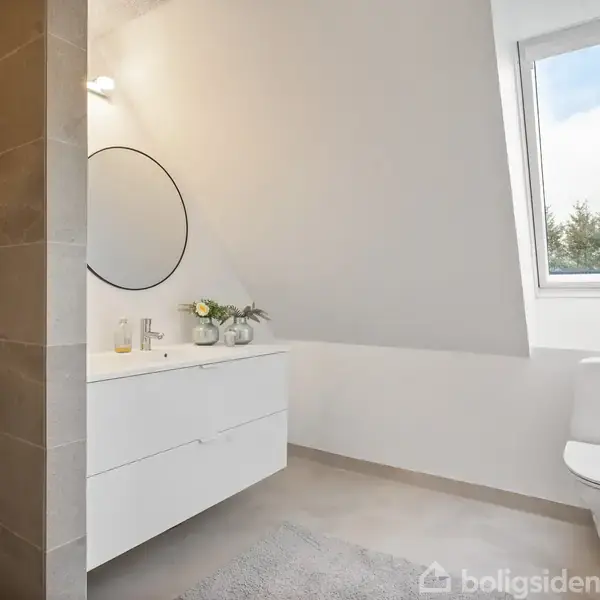Badeværelse med hvidt skab under rundt spejl. Til venstre ses en bruseniche. Foran er et vindue med udsigt til træer. Gulvet er gråt, og der er en toiletkumme.