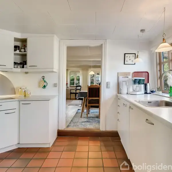 Køkken med hvide skabe og skuffer, hvor en kaffemaskine står på bordpladen under en lampe. Store vinduer giver udsigt til haven. Døråbning fører til spiseområde.