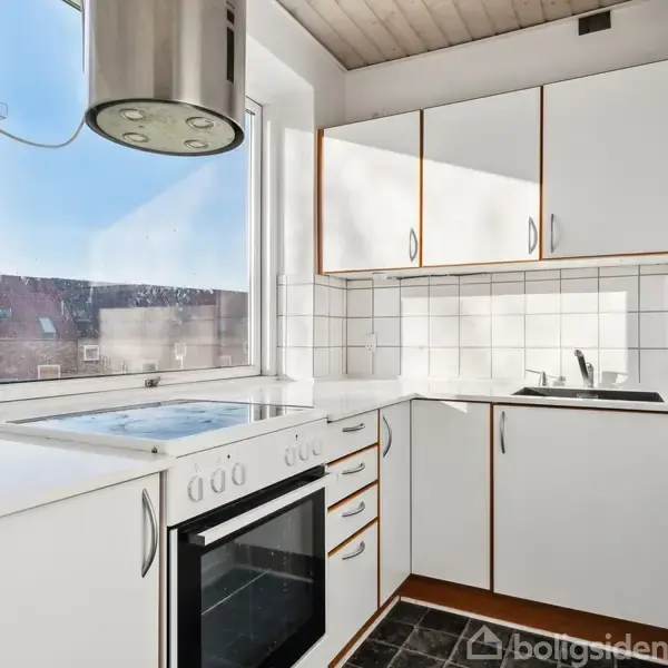 Et kompakt køkken med hvide skabe og fliser. En indbygget ovn under en kogeplade står ved et stort vindue, der giver udsigt til hustage. En vask ses til højre.