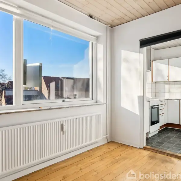 Et rum med trægulv og store vinduer der lukker sollys ind; til højre ses en døråbning til et køkken med hvide fliser og et komfur.