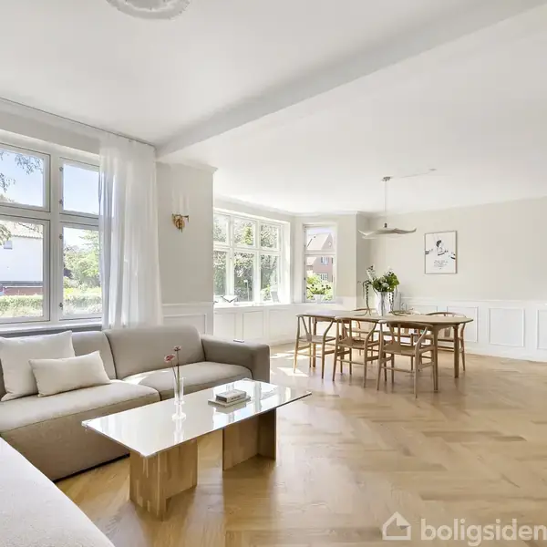 En lys stue med et hjørnesofaarrangement og sofabord ved vinduer. Et spisebord står midt i rummet, mens en kommode og dekorativ vase ses i baggrunden.