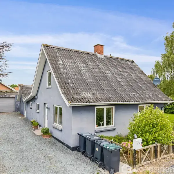Grå hus med spidst tag står stille ved en indkørsel af grus. I forgrunden er der affaldscontainere, mens grønne planter omgiver huset.