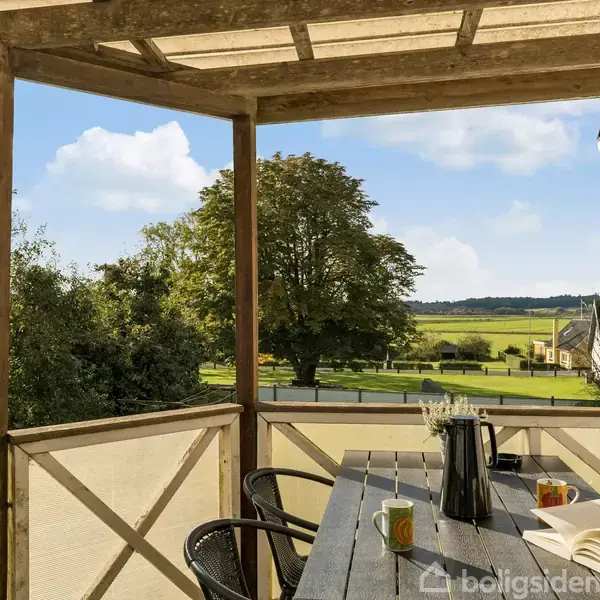 En træterrasse med bord og fire stole står i fokus. På bordet er der en kaffekande, et åbent magasin, og kopper. Terrassen har udsigt over en landlig, grøn natur.