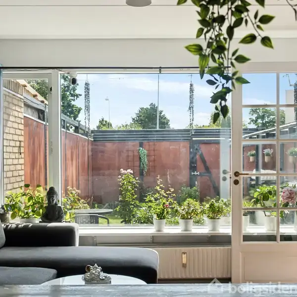 Sofa står i stue ved stort glasparti og fransk dør, åbner ud til en terrasse med blomster og planter. Sollys strømmer ind i det hjemlige rum.