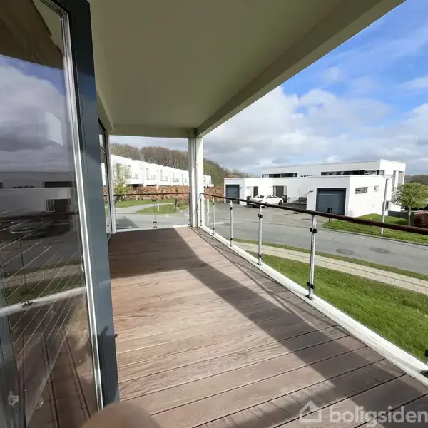 En rummelig altan med træplanker og glasrækværk strækker sig ud fra en moderne bygning. Udsigt over en stille, beboelsesvej med andre moderne huse og grønne plæner.