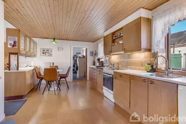 Et rummeligt køkken med træloft og brune skabe har en spiseplads med fire brune stole. Køkkenet er veludstyret med ovn, vask, og vindue med blondegardin.