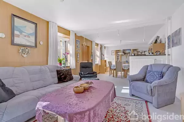 En stue med en grå sofa og lænestol omkranset af lyse tæpper på gulvet; et bord med en lilla dug i midten. Bagved er der en spiseplads med seks stole.