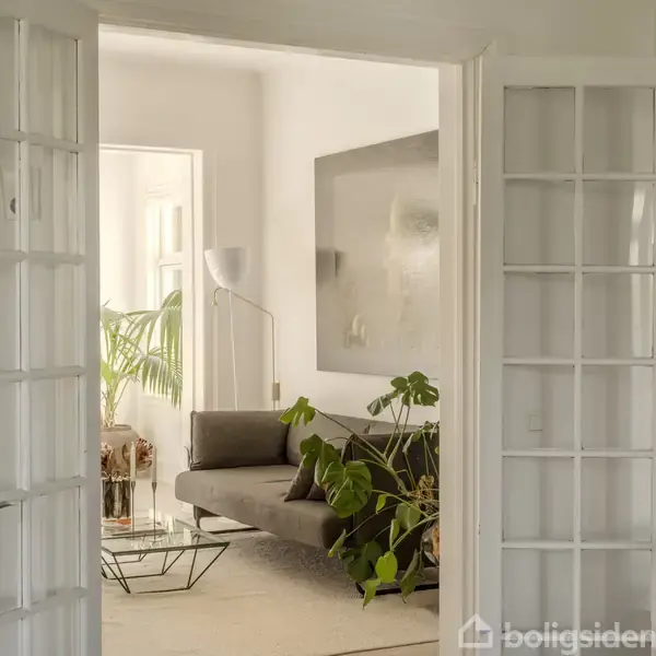 Dobbelt glasdør åbner til en moderne stue med en grå sofa, stor stueplante og kunst på væggen. En gulvlampe står ved vinduet ved siden af en plante.