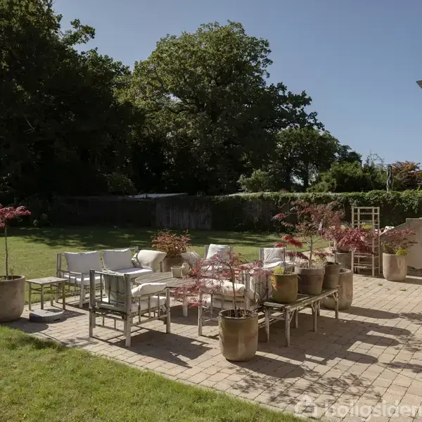 Terrassemøbler står arrangeret på en flisebelagt terrasse med potteplanter omkring. I baggrunden ses en græsplæne omkranset af træer og buske i en solrig have.