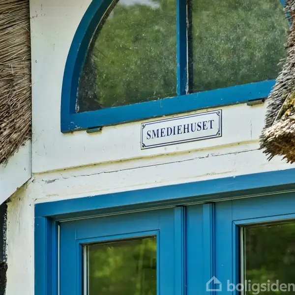 Blå hoveddør under halvbuet vindue; bygningen har stråtag. Skilt over døren: "SMEDIEHUSET". Omgivelserne indikerer en traditionel, rustik byggestil.