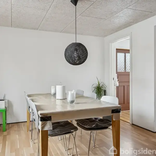 Et spisebord dækket med dug står i midten af et rum. Over bordet hænger en rund loftlampe. Rummet har lyse vægge, trægulv og en grøn plante i baggrunden.