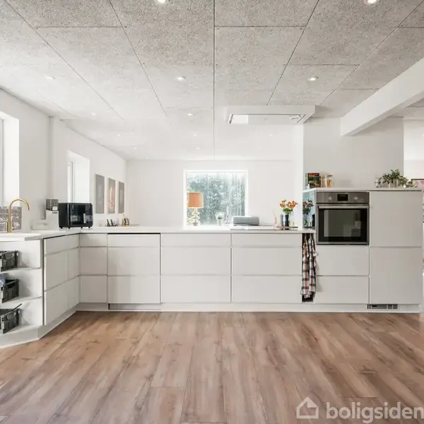 Et moderne køkken med hvide skabe og trælaminatgulv står stille i sollys, omgivet af åbne rum og vinduer, der giver udsigt til natur udenfor.