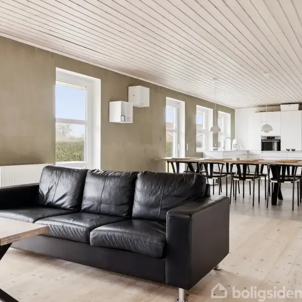 Sort lædersofa står i stue med træloft og trægulv, mod et spisebord med sorte stole. Store vinduer i rummets længde giver naturligt lysindfald. Ingen tekst til stede.