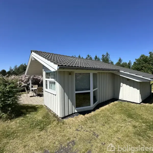 Hus med grå facade og sort tag. Det står på en velplejet græsplæne omgivet af træer og buske. En terrasse med havemøbler ses ved siden af huset.