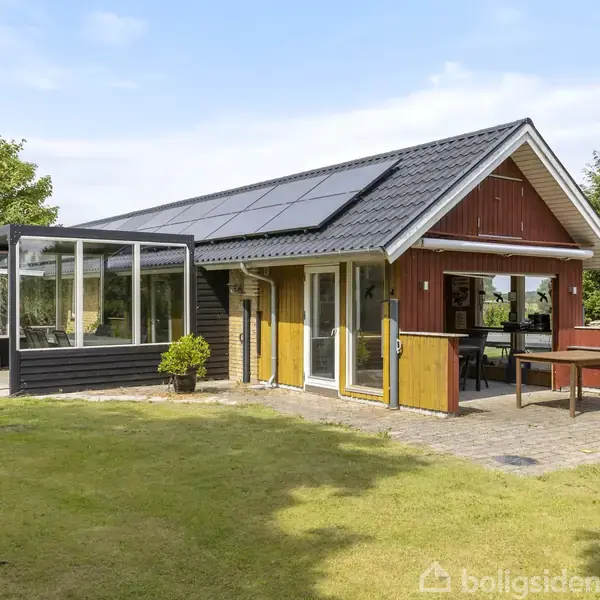Et lille træhus med solpaneler på taget står i en grøn have. Huset har en tilstødende glaskonstruktion og udendørs terrasse omgivet af træer.