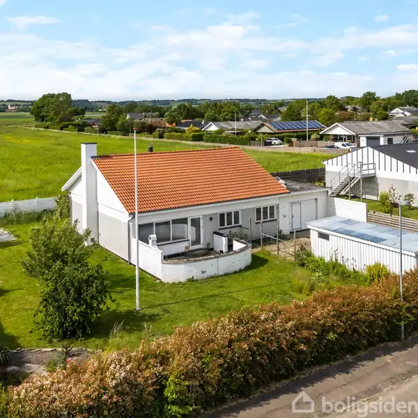 Et hvidt hus med rødt tag står i en grøn have med flagstang. I baggrunden ses en landlig scene med flere huse og marker under en blå himmel.
