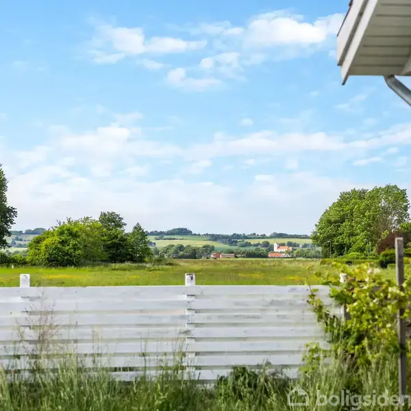 Husdetalje; sidebeklædning og tagrende ved hvidt hegn; baggrund med grønne marker og træer under en blå skyet himmel.