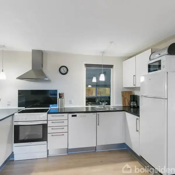 Et moderne køkken med hvidt inventar har en ovn og emhætte. Et køleskab står ved siden af en mikroovn. To vinduer og lamper giver lys over den åbne arbejdsplads.