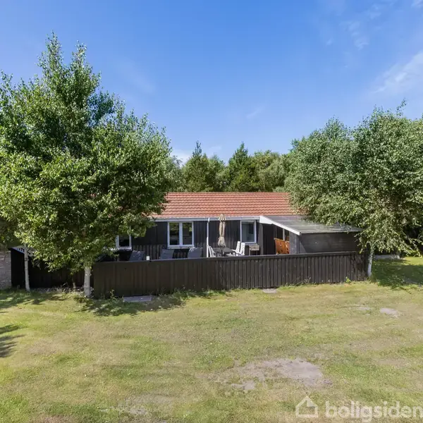 Et sort træhus med rødt tag står blandt træer. Udendørs terrasse med havemøbler. Omkringliggende græsplæne og skov giver en naturrig atmosfære.