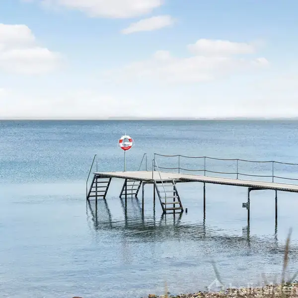 En badebro strækker sig ud over stille vand med en redningskrans på gelænderet. Baggrunden har en klar, lys himmel og en synlig horisont over havet.