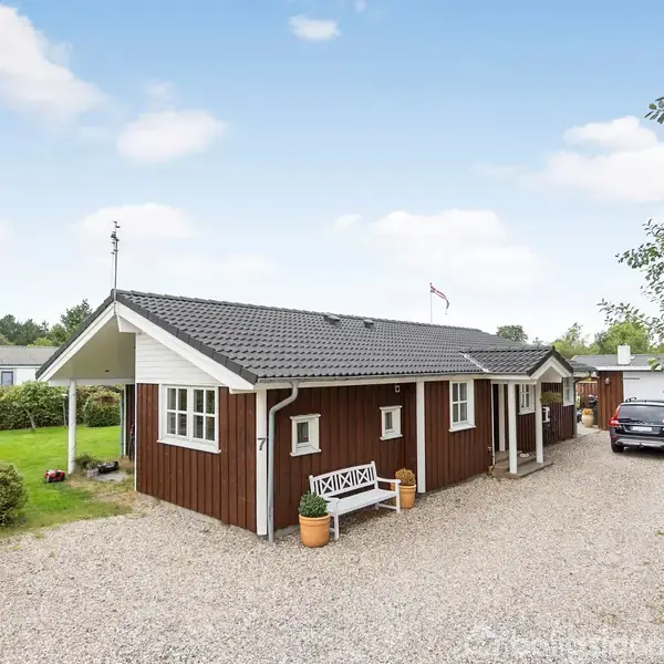 Et træhus står stille i en have med grusvej og parkeret bil, omgivet af grønne buske og træer. Nummer 7 ses på huset, der har en lille bænk foran.