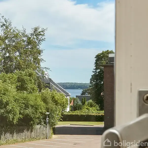 Vindueshåndtag på en åben dør med udsigt over en gade med træer og hegn. I baggrunden ses et hustag og en flod eller sø under en blå himmel.
