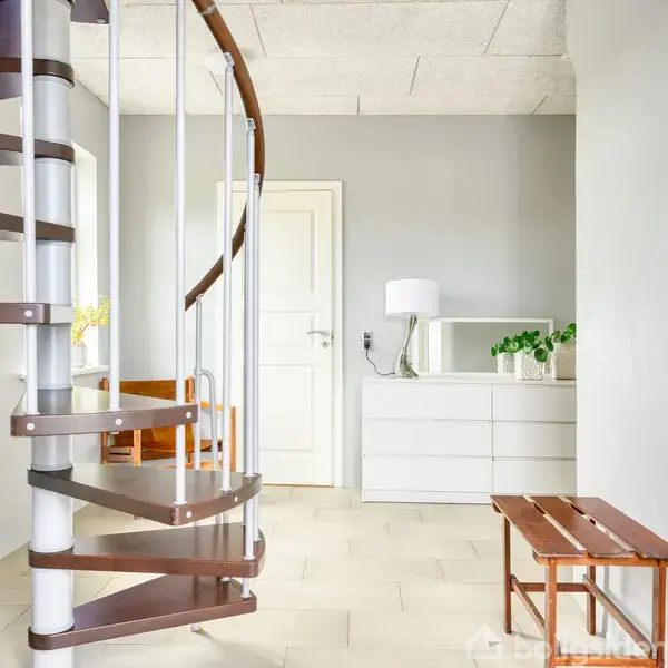 En spiraltrappe af mørkt træ står i et lyst rum ved siden af en hvid kommode med en lampe og planter; en bænk står i forgrunden.