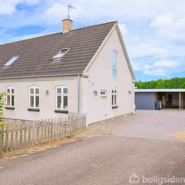 Et hvidt, muret hus med mørkt tag står i et åbent område. Til højre for huset er der en carport. Grusbelagt indkørsel og træhegn foran huset.