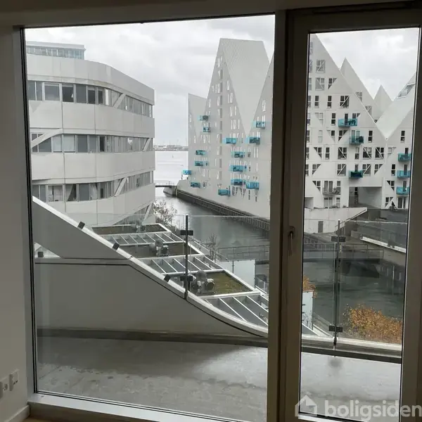 En lys stue med en beige sofa til venstre, en TV-skærm på et hvidt skab til højre, og en udsigt til moderne bygninger gennem et stort vindue foran.
