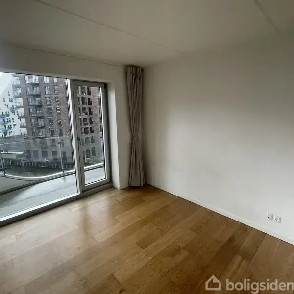 Et sæt lysegrønne havemøbler står på en altan med glasrækværk. I baggrunden ses moderne murstensbygninger og en lille kanal omgivet af træer i et bymiljø.