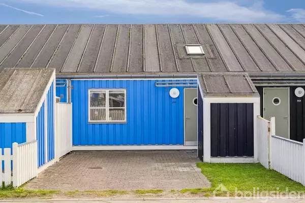 Blåt hus med et skifertag og et enkelt vindue foran, omgivet af en hvid stakit og en flisebelagt indkørsel. Himlen er klar med få skyer.