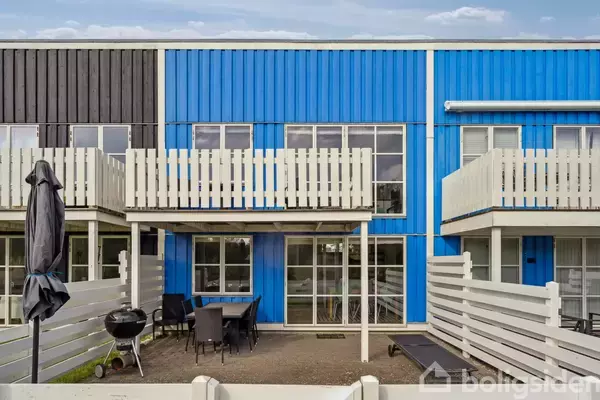 Bagside af et moderne rækkehus med blå facade og hvide altaner. I gårdhaven er der et bord med stole og en grill, omgivet af et hegn.