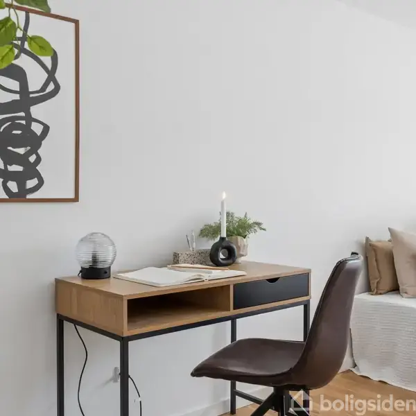 Et skrivebord står i et lyst rum med en stol foran sig. Der er en tændt lampe og en åben bog på bordet. I baggrunden ses en seng med puder.
