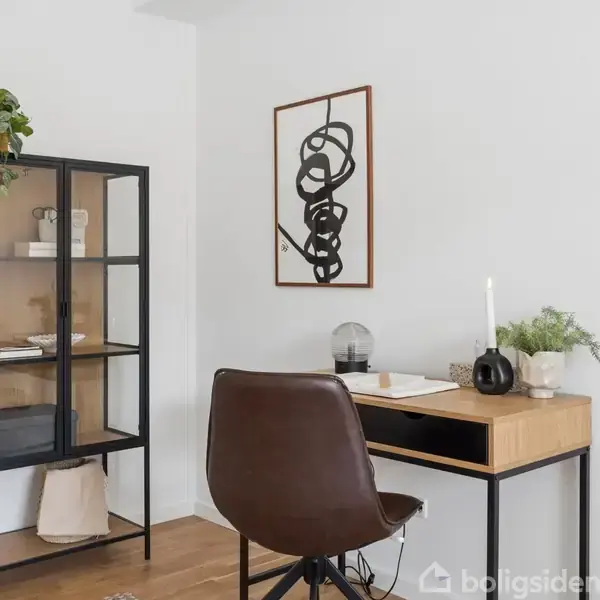 Et skrivebord med en brun stol står ved siden af en glasmontre med bøger og planter. En kunstplakat hænger på væggen. Rummet har lyse vægge og trægulv.