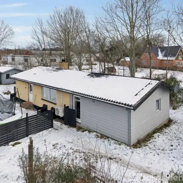 En enkeltstående gul og grå bygning er dækket af sne. Den er omgivet af hegn og træer i et snedækket landskab. En trampolin ses ved siden af huset.