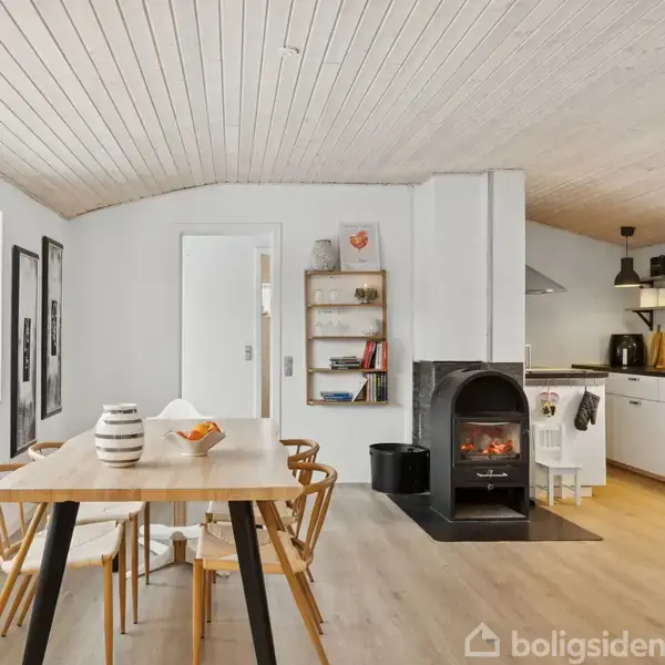 Spisebord med stole står i et lyst, moderne køkken-alrum med træloft, hvor en brændeovn er placeret ved siden af et åbent køkkenområde med hvide skabe og sorte hylder.