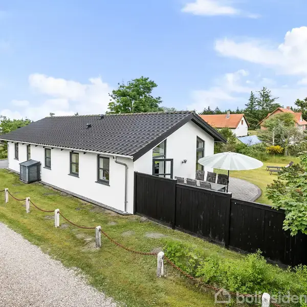 En hvid murstenshus med sort tag står i en grøn have. Til venstre findes en grussti, mens der er en terrasse med parasol til højre.