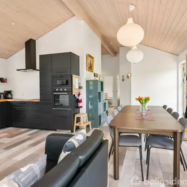 En moderne køkken-stue med sorte skabe og træloft. Et spisebord med blomster står i midten, omgivet af stole. Store vinduer lader naturligt lys strømme ind.