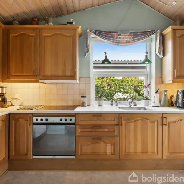 Køkken med træskabe og hvidevarer. En kaffemaskine og mikroovn står på bordpladen. Et vindue med udsigt til en have og to hængelamper over vasken.