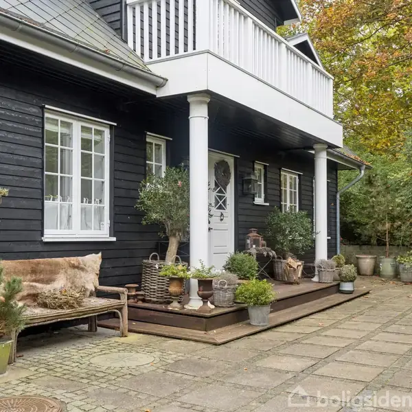 Et sortmalet hus med hvide vinduer og dør står ved en stenterrasse. Planter i krukker pryder området, og en bænk står ved siden af døren. Træer omkranser stedet.