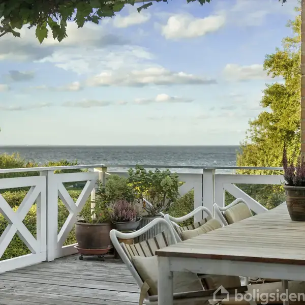 Terrasse med et bord og stole, står med udsigt over havet. Omgivet af grønne planter og en hvid rækværk. Himlen er let skyet.