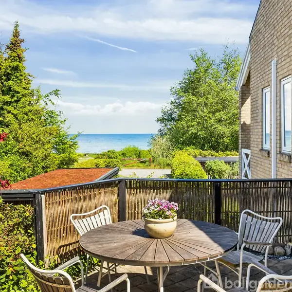 Rundt havebord med fem stole står på en terrasse, prydet med en blomsterkrukke. Omgivet af grønne planter med udsigt til havet i baggrunden ved siden af et hus.