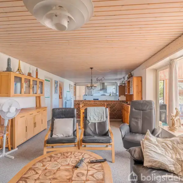 Stue med lænestole og sofabord i forgrunden, set mod køkkenet med træmøbler langs væggene. Store vinduer på højre side lader naturligt lys komme ind i rummet.